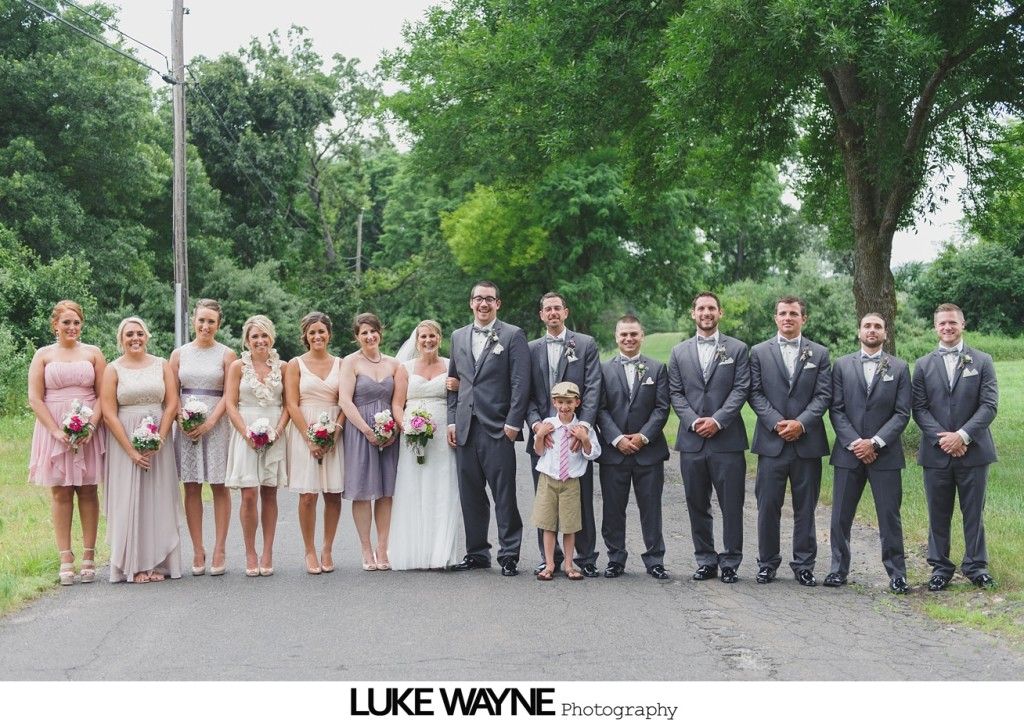 Wedding party posing on a rural road; bride and bridesmaids in pastel dresses, groomsmen in gray suits.