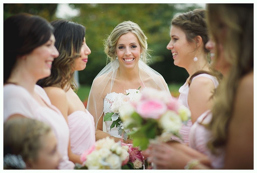 Bride smiles surrounded by bridesmaids in pink dresses, holding bouquets, outdoors.