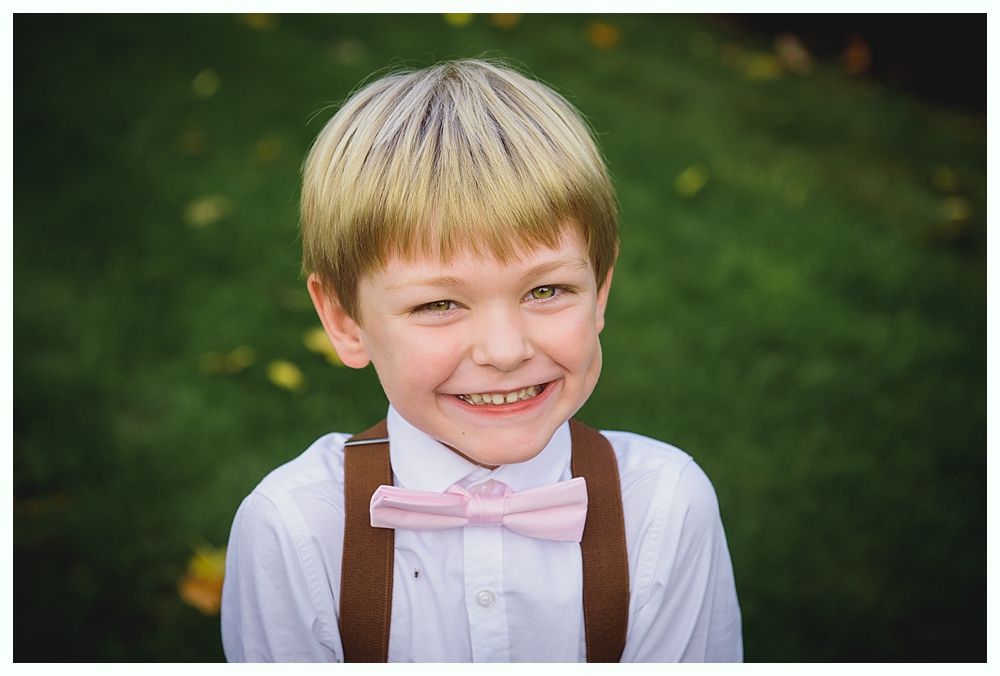 Boy smiling, wearing a pink bow tie and suspenders. Green background.