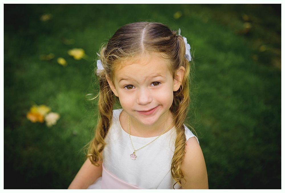Young girl with pigtails, wearing a white dress and smiling, in a grassy outdoor setting.