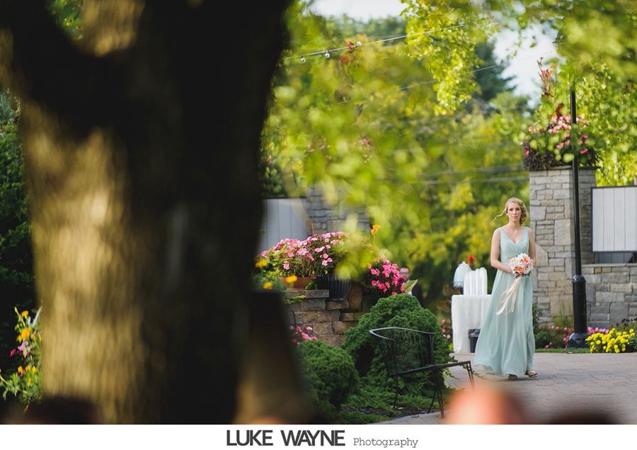 Bridesmaid in a light blue dress walks down the aisle, holding flowers. Outdoor setting with greenery and flowers.