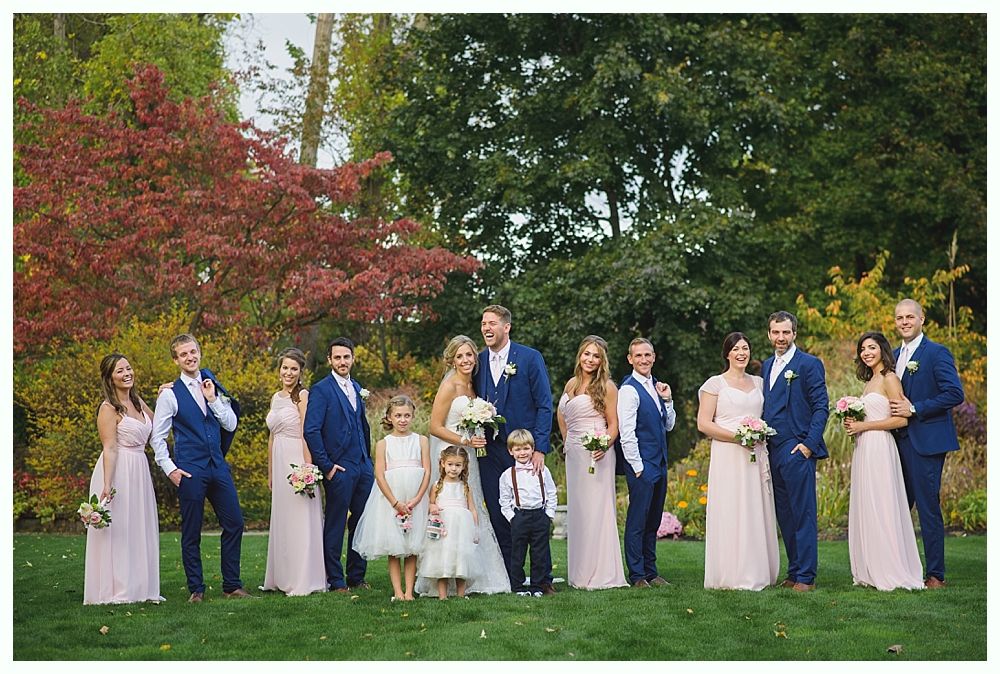 Wedding party on a lawn; couple in the center, bridesmaids in pink, groomsmen in blue suits, colorful fall foliage.