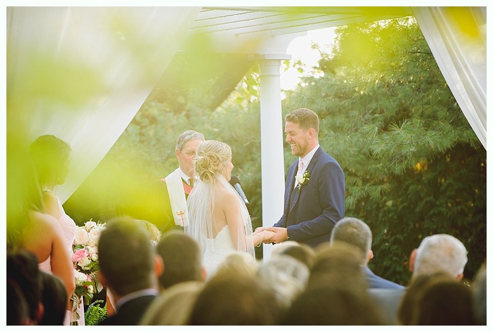 Bride and groom exchanging rings under a gazebo during an outdoor wedding ceremony; guests in attendance.