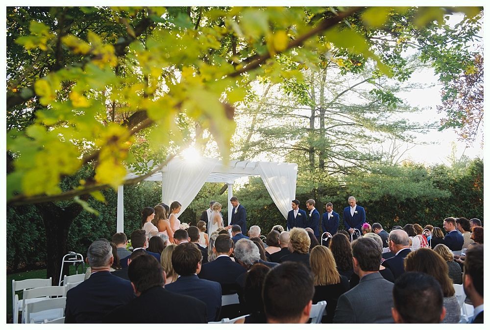 Wedding ceremony under a white canopy; guests seated, bridesmaids and groomsmen stand with the couple.