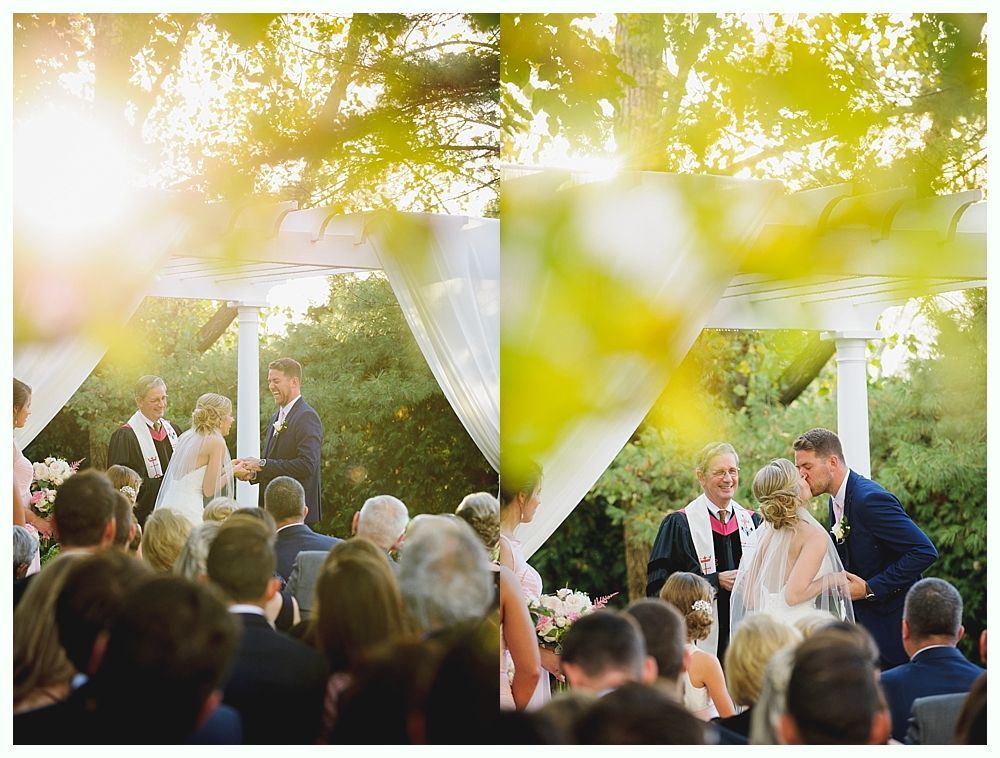 Wedding ceremony with couple kissing under white arbor in sunny outdoor setting.