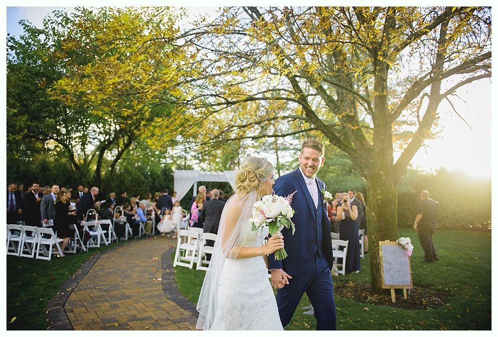 Newly married couple walks down aisle after outdoor ceremony. Guests cheer.