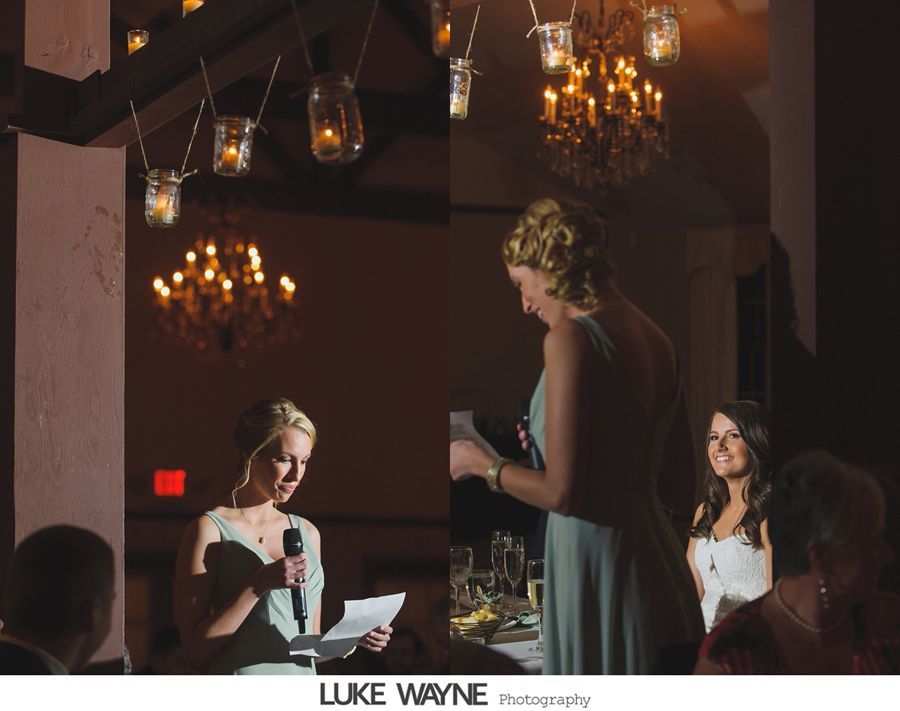 Woman giving a speech at a wedding, in a warmly lit room with hanging candle jars and chandeliers.