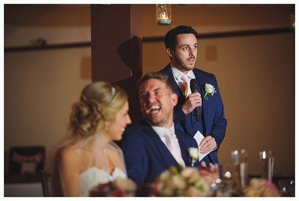 Man in blue suit giving a wedding speech; bride smiles, man in suit laughs. Indoor setting, warm lighting.