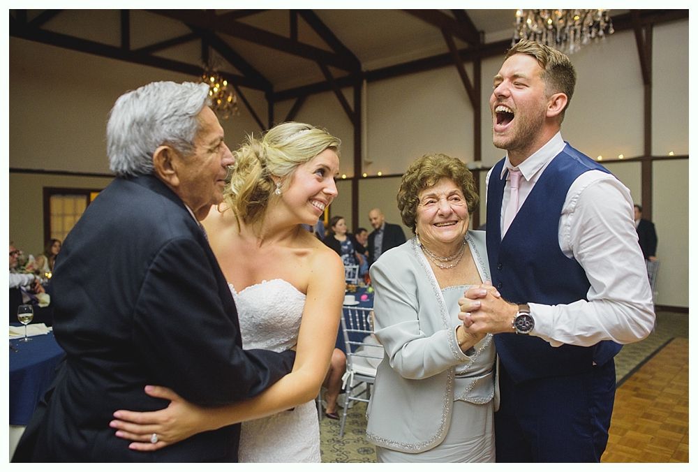 Bride and groom dance with family at a wedding reception; happy faces, wooden interior.