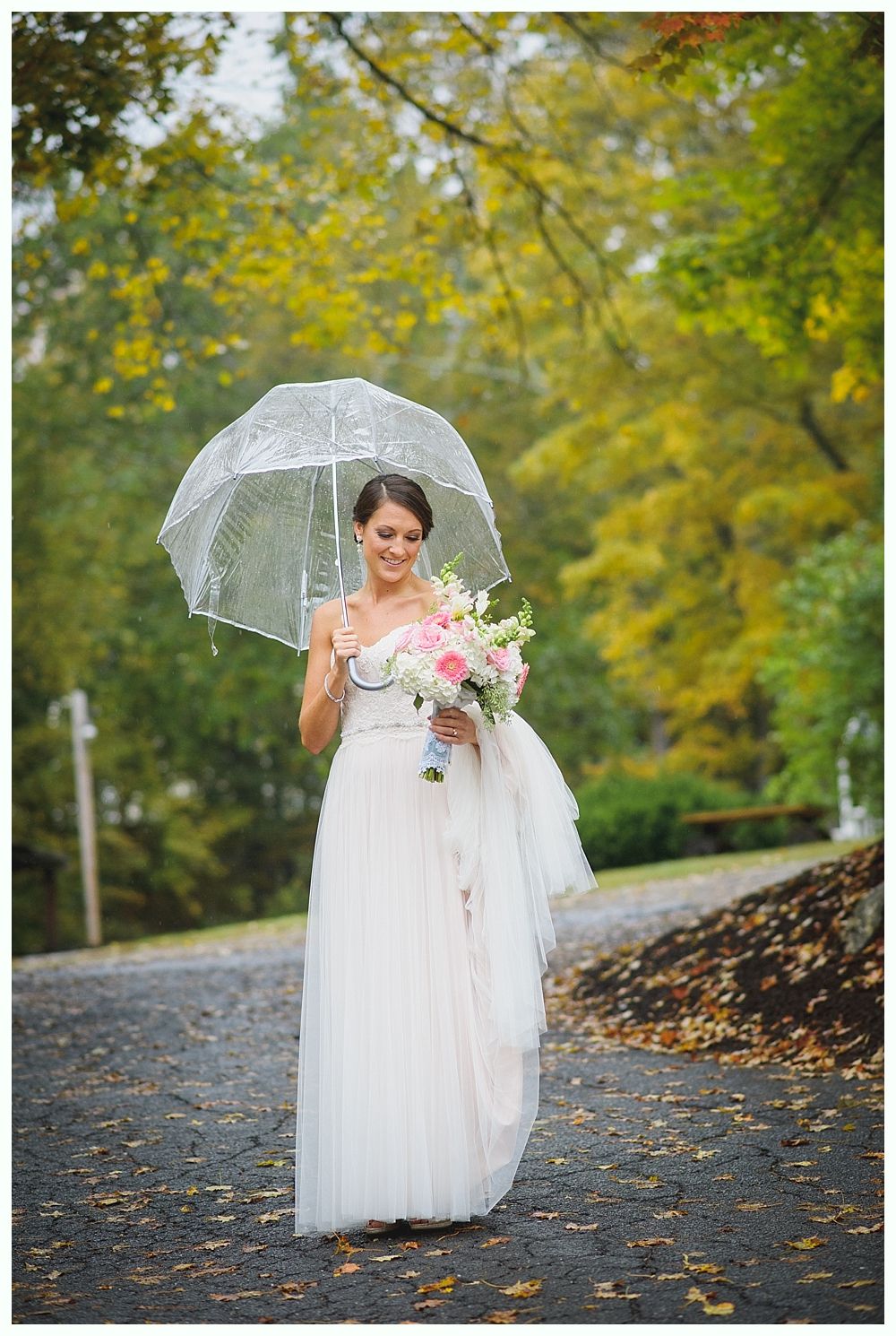 Bride in white dress holds clear umbrella and bouquet, standing on a path with fall foliage.