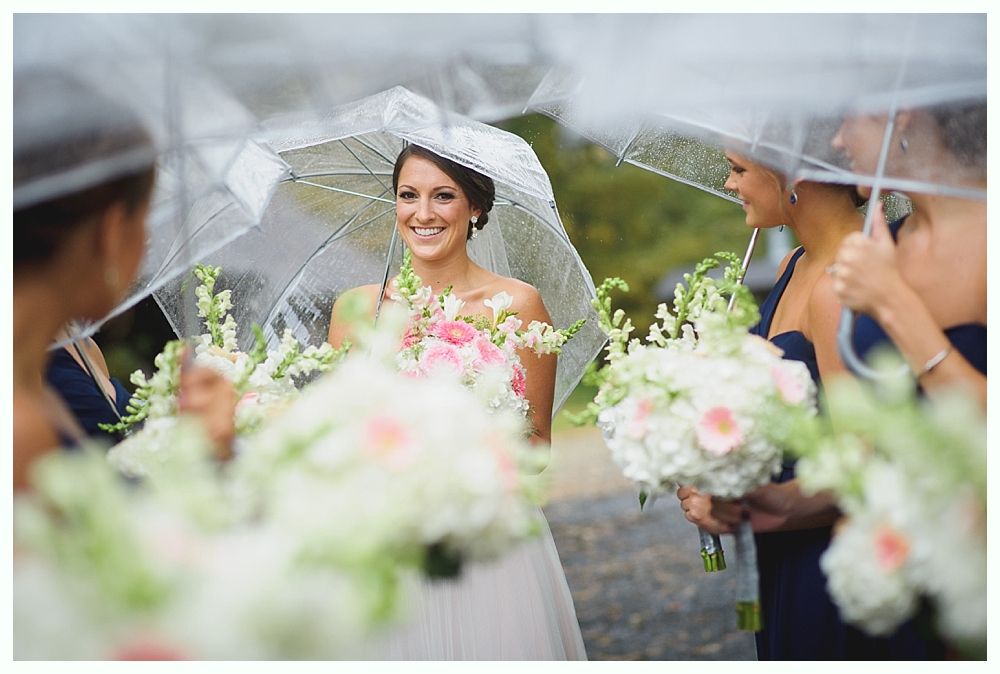 Bride smiling under a clear umbrella, surrounded by bridesmaids holding bouquets in the rain.