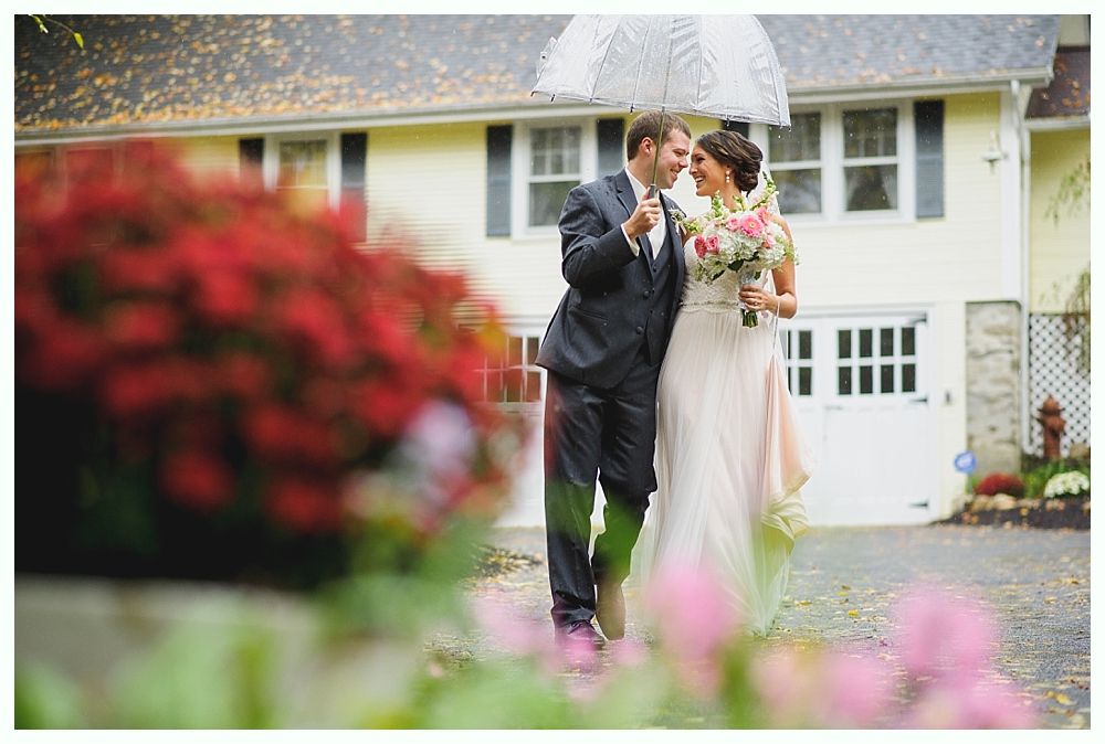 Bride and groom under umbrella in front of a yellow house, holding hands and smiling.
