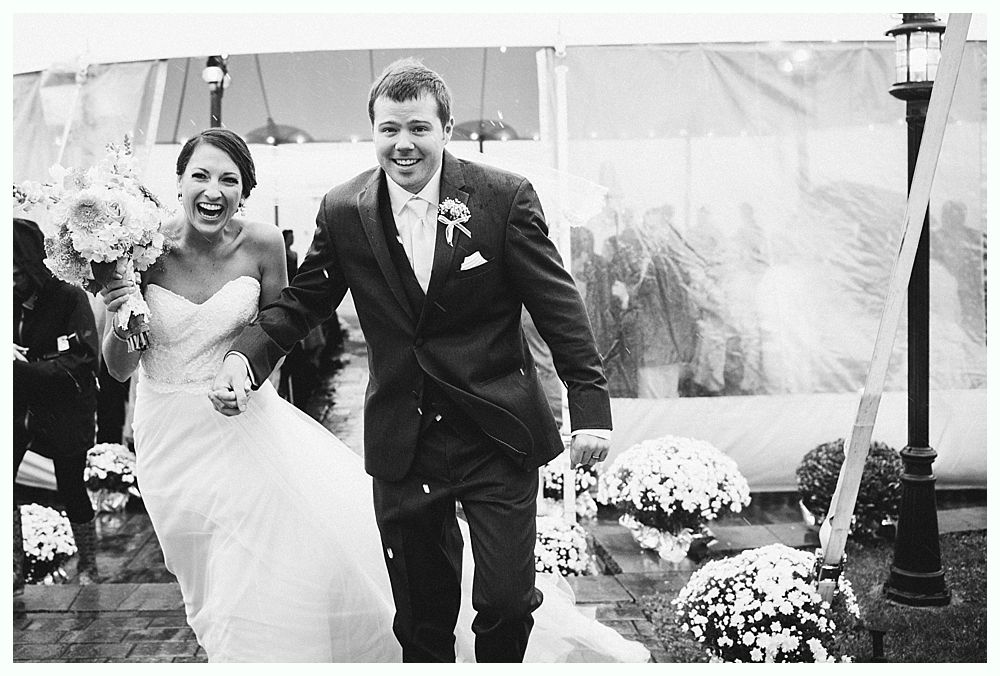 Bride and groom exit a wedding ceremony, smiling. She wears a white gown, he wears a suit. Flowers surround them.