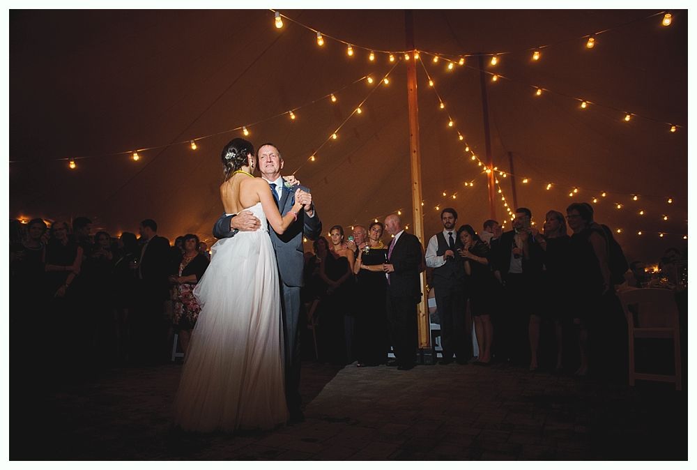 Bride and groom dance under string lights at wedding reception; guests watch.