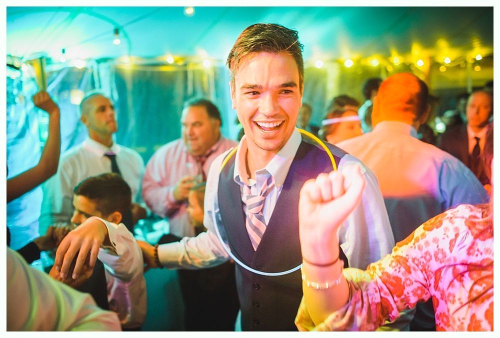 Man dancing with a glowing necklace, smiling in a party setting with people in the background.