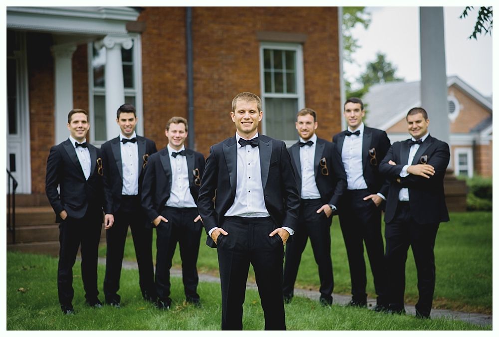 Men in black tuxedos pose for a photo in front of a brick building.