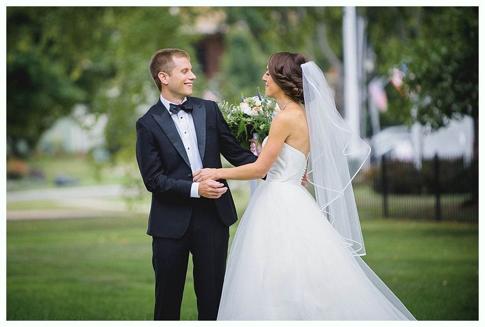 Bride and groom smiling at each other, holding hands, outdoors in a park, bride in white dress, groom in tuxedo.