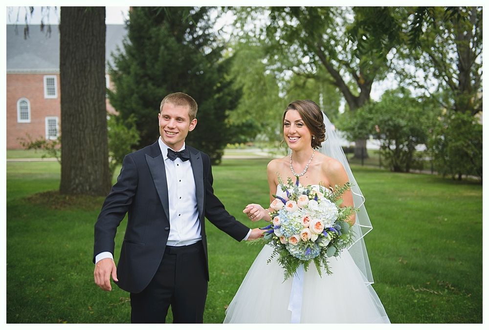 Wedding couple holding hands, smiling, in a grassy area with trees and a building in the background.