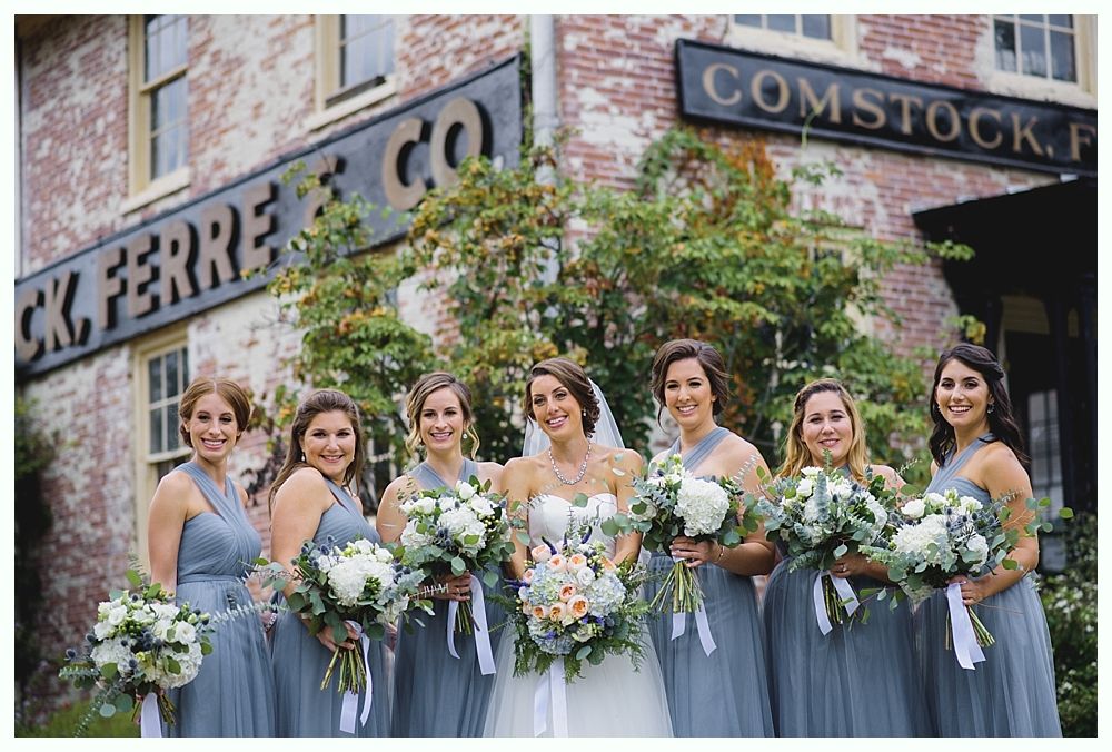 Bride with bridesmaids holding bouquets in front of brick building with sign. All in light blue dresses.