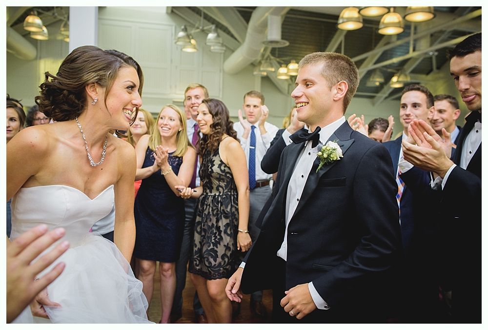 Bride and groom smiling at each other during a wedding reception, surrounded by guests clapping.