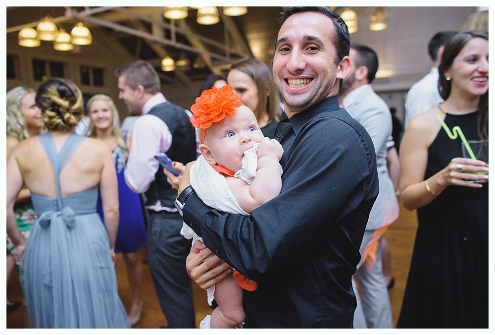 Man holding baby, both smiling at a party. Baby wears an orange flower headband.