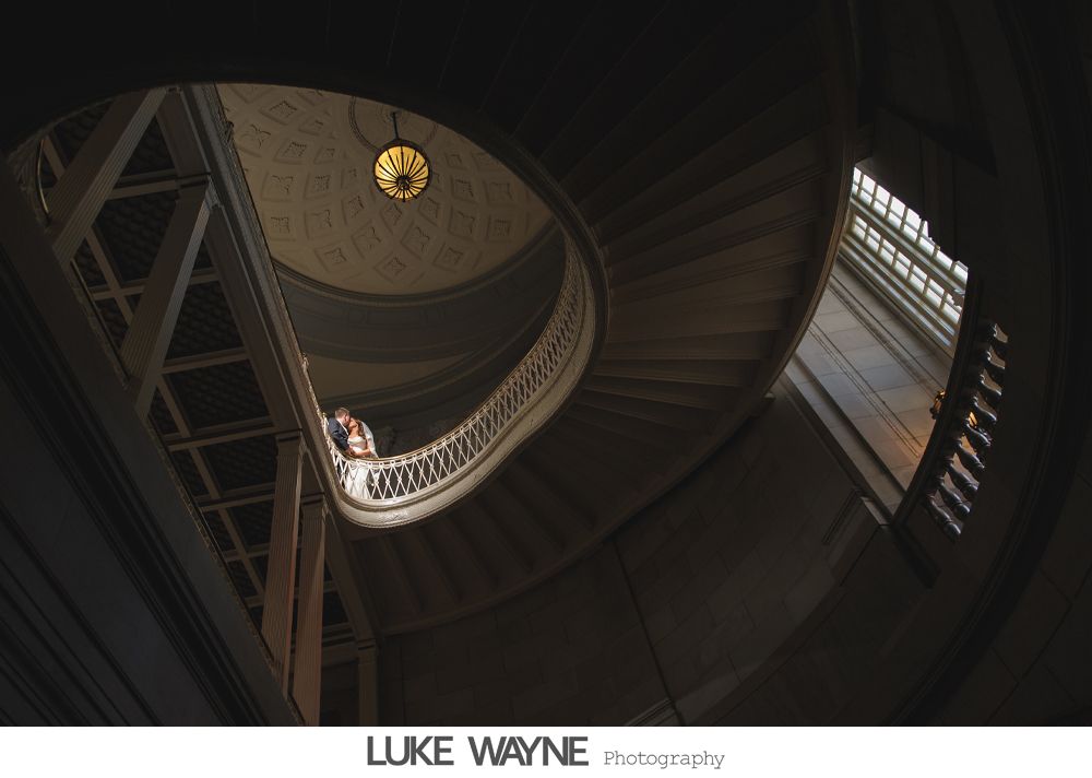 Spiral staircase, couple on the railing, bathed in light, inside a grand building.