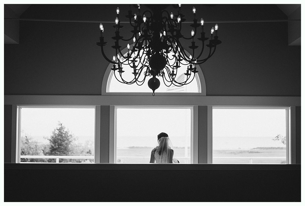 Woman silhouetted by large windows, looking toward the horizon. Chandelier hangs above.