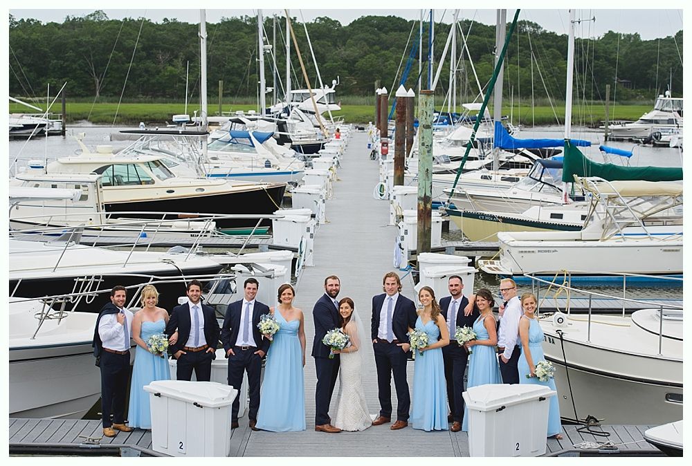 Wedding party posing on a dock with boats; bride and groom in center, bridesmaids in blue dresses, groomsmen in suits.