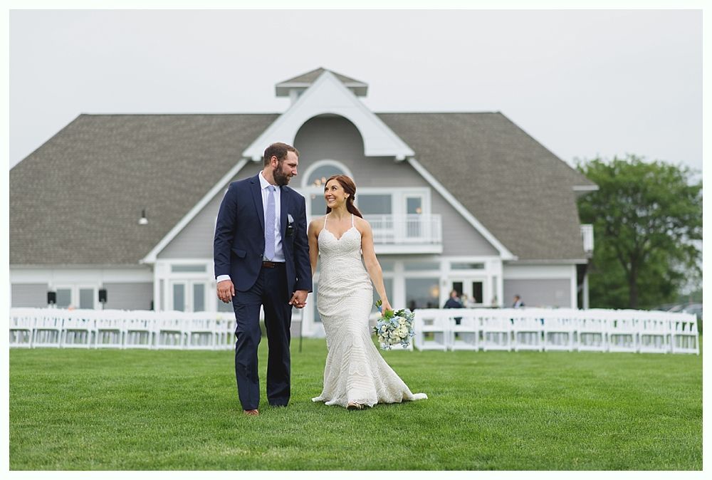 Bride and groom walk hand-in-hand on grass, smiling. Building with grey roof in background.