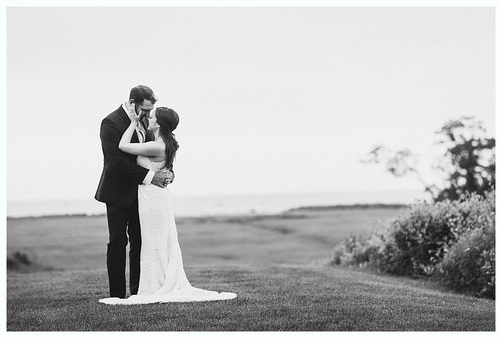 Couple embraces on a grassy hill overlooking a body of water; wedding attire.