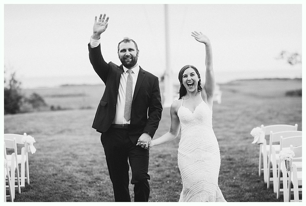 Newlyweds waving, holding hands, smiling outdoors after ceremony.