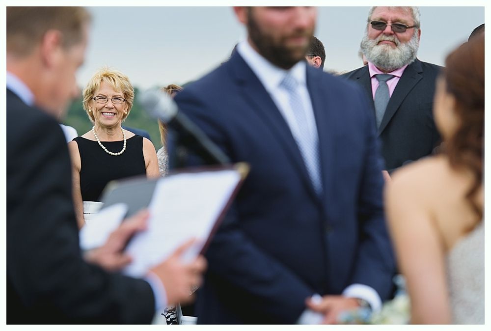 Guests smiling during an outdoor wedding ceremony; officiant reading; groom and bride in focus.