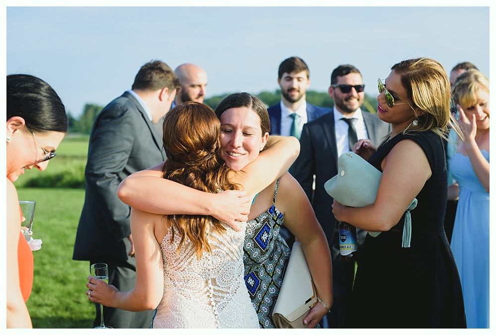 Two women embrace outdoors, surrounded by guests. People smile, some wear formal attire.