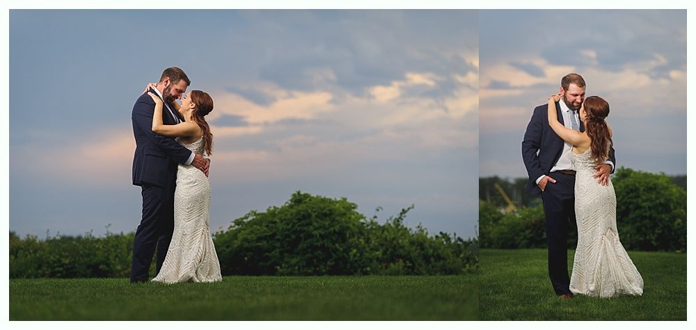 A newly married couple embraces on a grassy field with a cloudy sky backdrop.