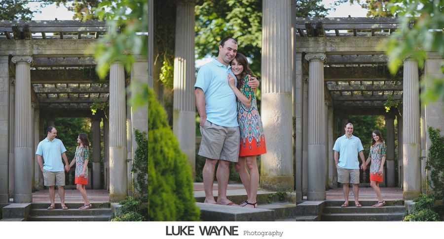 Couple poses in stone archway, framed by columns and greenery. Man in blue shirt, woman in patterned dress.