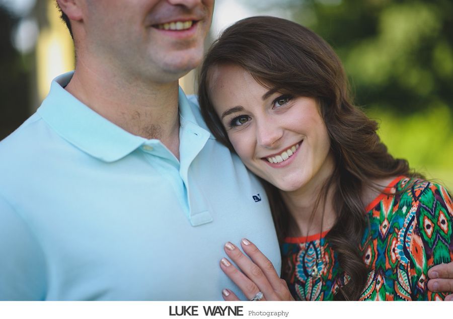 Woman resting her head on a man's chest; smiling, outdoors, in front of greenery.