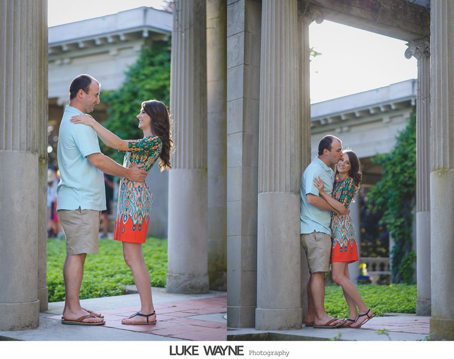Couple embracing near stone columns in garden setting. Man in light blue shirt and shorts, woman in patterned dress.