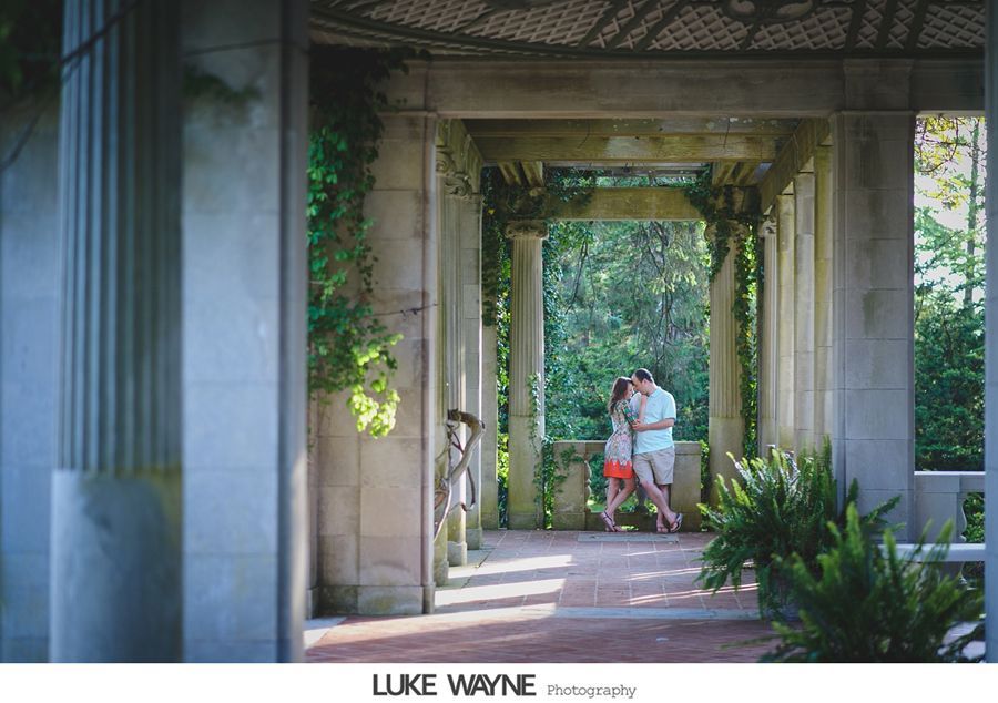 Couple kissing in a stone archway with greenery and columns, warm light, brick floor.