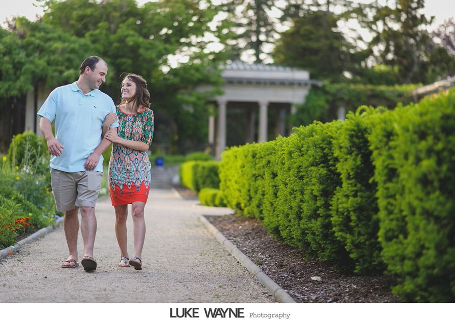 Couple walking hand-in-hand in a garden with green hedges and a stone path. Man in blue shirt, woman in patterned dress.