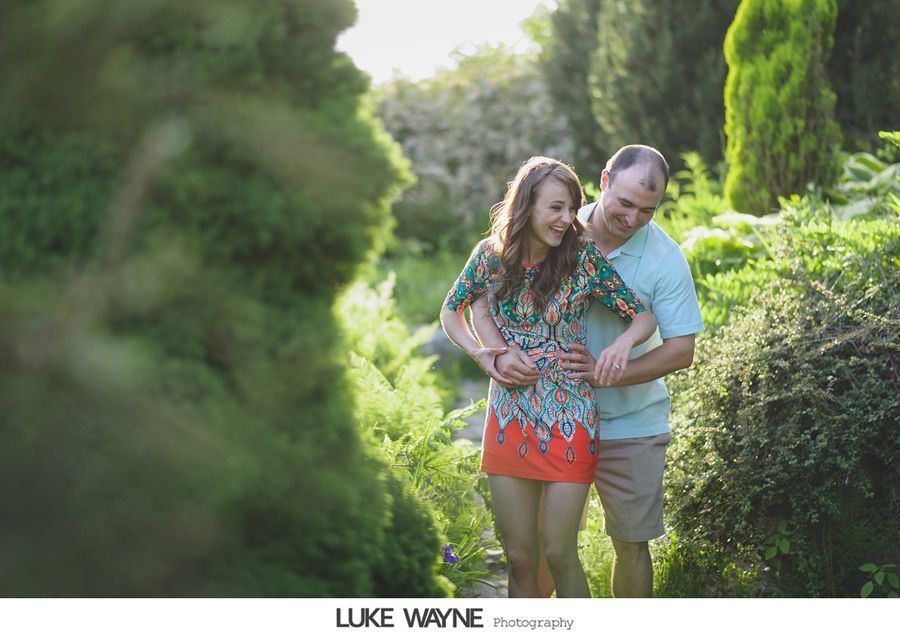 Couple embraces in a lush garden, woman laughs, man smiles, both in casual attire.