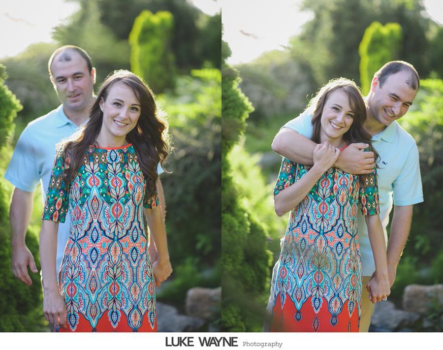 Couple posing outdoors; man embraces woman from behind, smiles, trees in background.