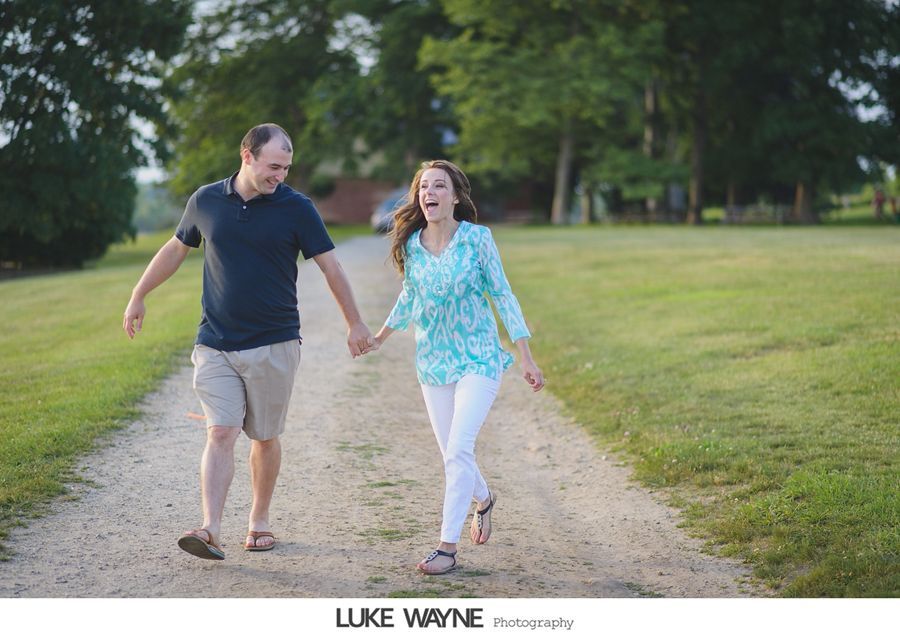 Couple holds hands and runs down a dirt path, woman laughing, man smiling, green grass, trees in background.
