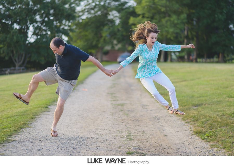 Couple holding hands, jumping on a gravel road. Woman in white pants, turquoise shirt. Man in khaki shorts, navy polo.