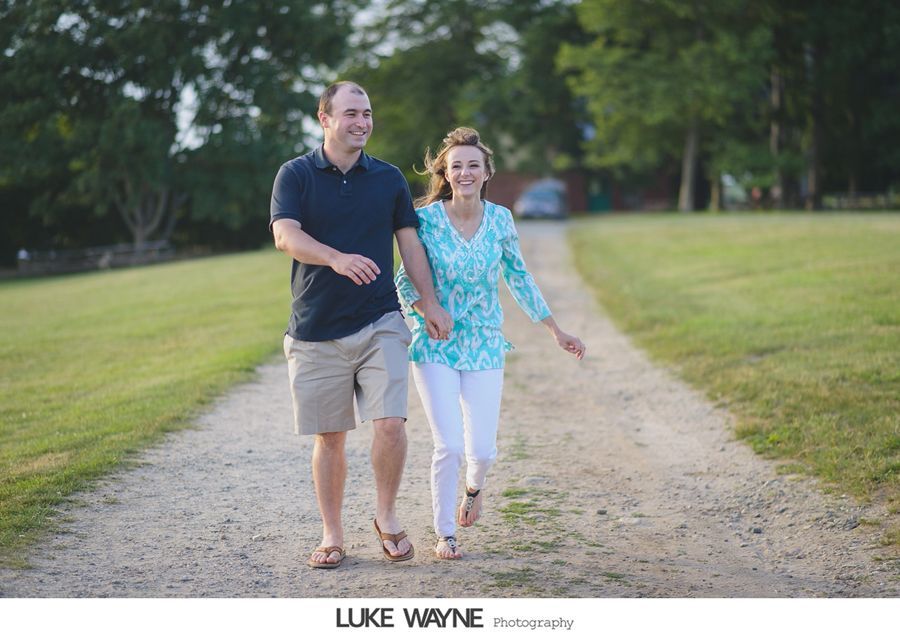 Couple walking and holding hands on a gravel path, laughing. Green grass and trees in background.