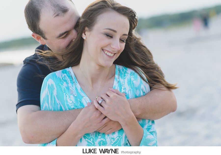 Man hugging woman from behind on a beach; woman smiles, ring visible, blue shirt.