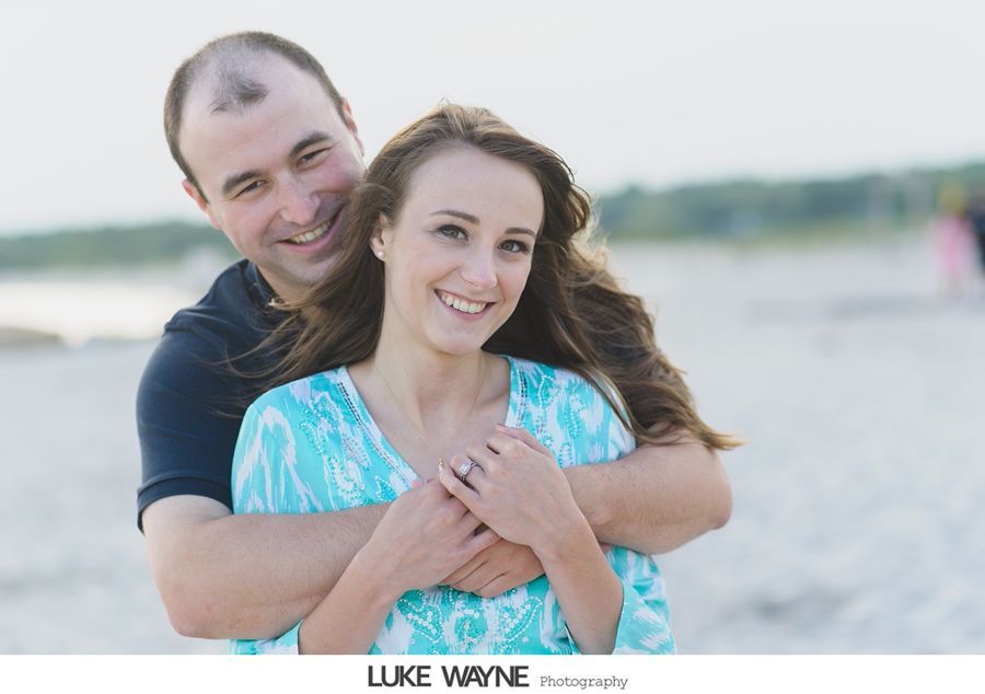 Man hugging woman from behind on a beach, both smiling. The woman shows off a ring.