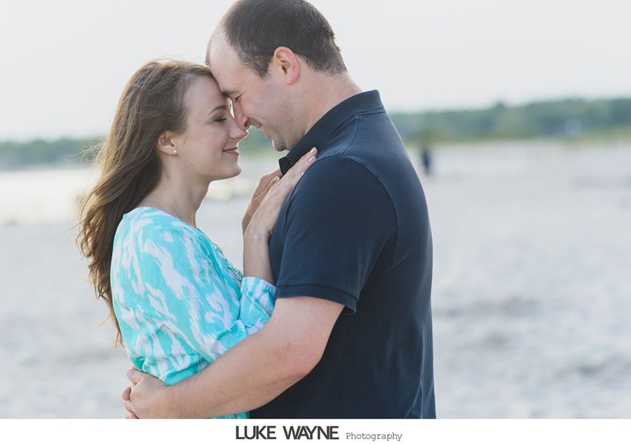 Couple embraces on a beach, faces close, looking at each other. Man in navy shirt, woman in turquoise top.