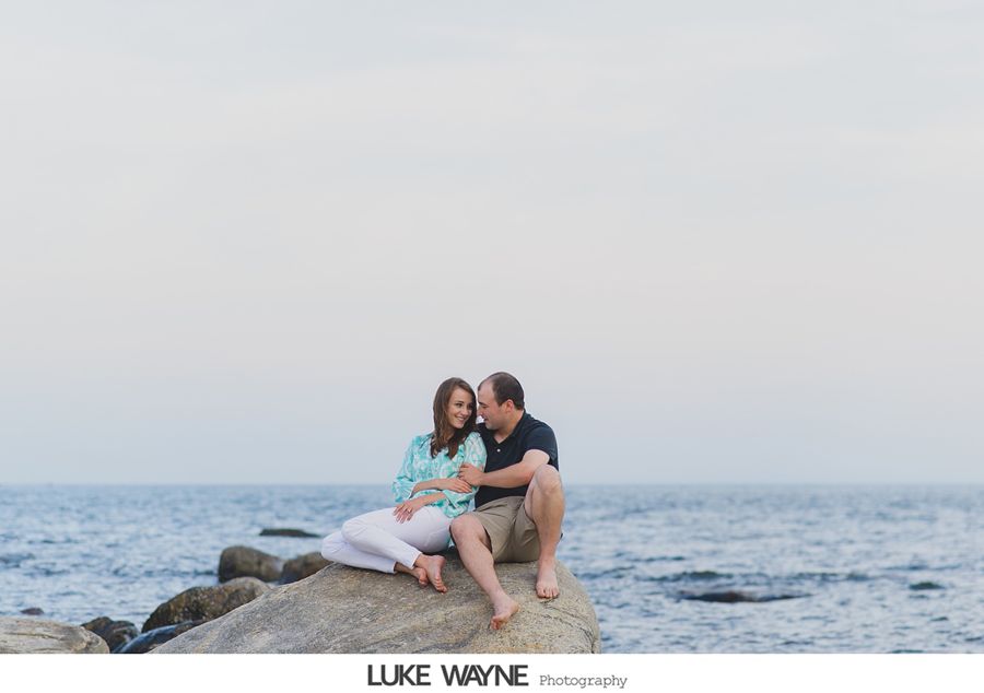 Couple sitting on a rock by the ocean, smiling and looking at each other.