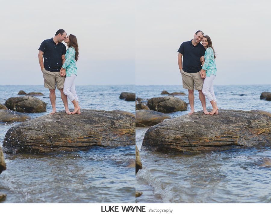 Couple embracing on a rock in the ocean. Man in black shirt, woman in teal. Blue water, clear sky.