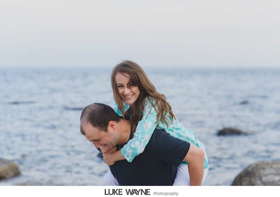 Man gives woman a piggyback ride on a beach, smiling. Ocean in background.